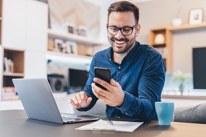 man smiling looking at phone with laptop in front of him